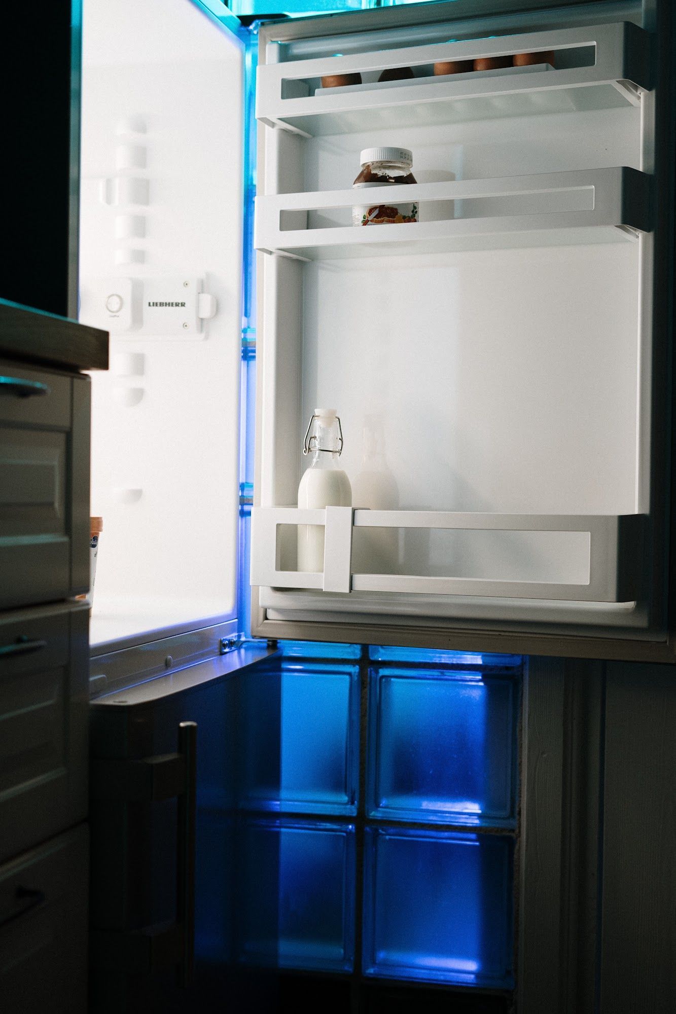 Open refrigerator interior, mostly empty. Milk bottle on middle shelf, blue light in drawers.