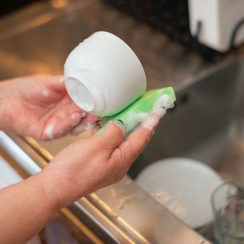 Hands washing a white cup with a green sponge in a kitchen sink.