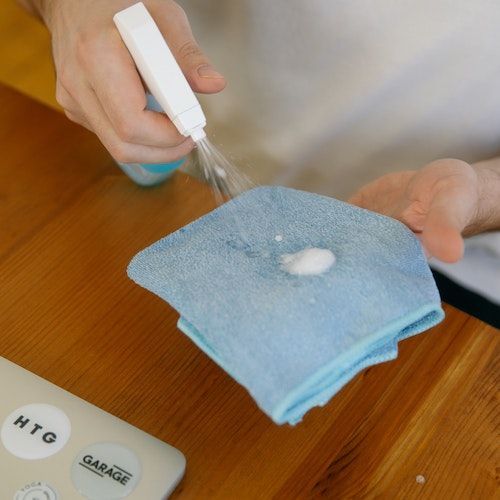 Person spraying cleaner onto a blue cloth on a wooden table. A laptop with stickers is visible.