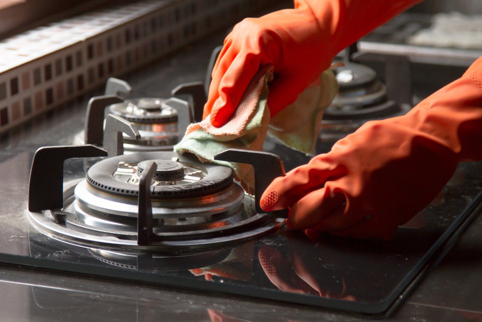 A person wearing orange gloves is cleaning a gas stove with a cloth.