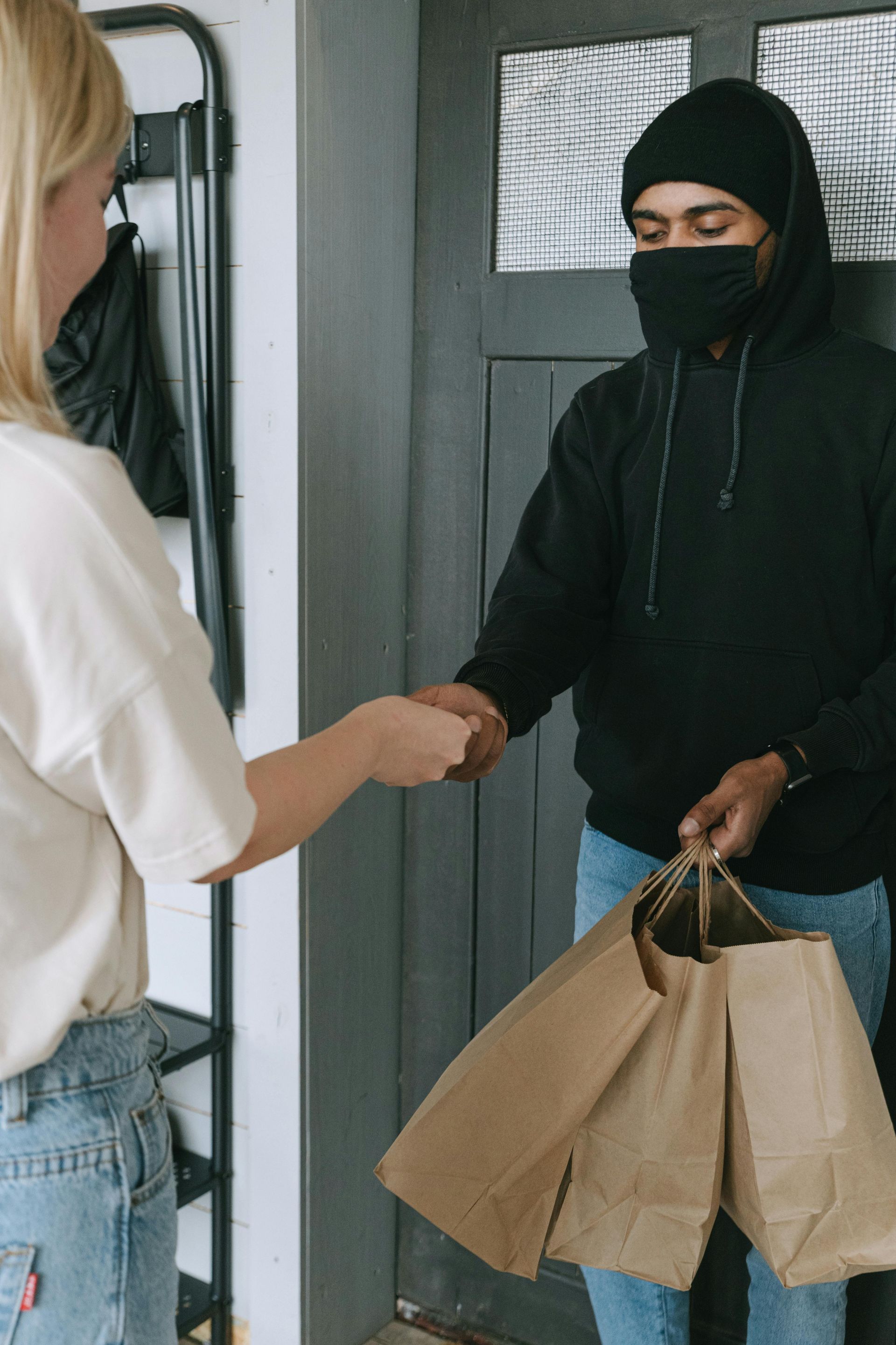 Two people exchange a paper bag at a doorway, one wearing a black mask and holding shopping bags.