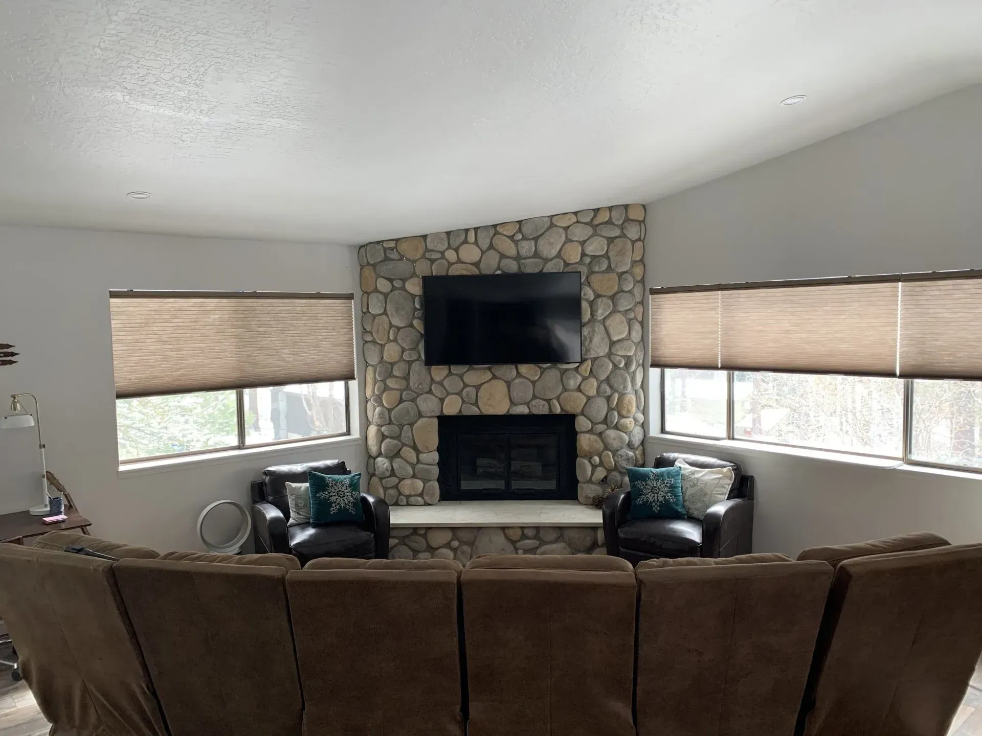 Living room with stone fireplace, wall-mounted TV, and two windows with blinds behind a brown couch