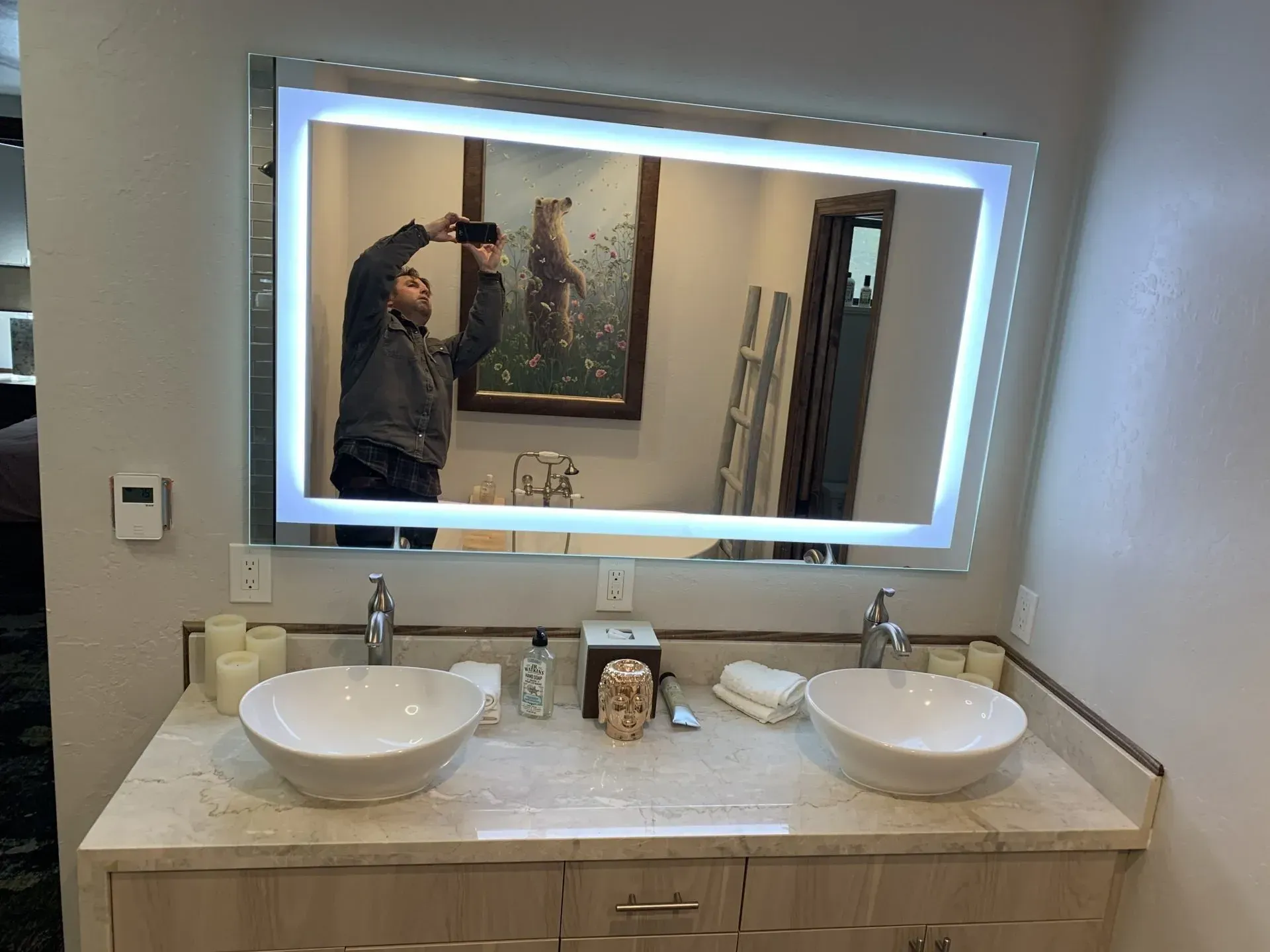 Bathroom vanity with two vessel sinks and illuminated mirror, reflecting a person taking a photo.