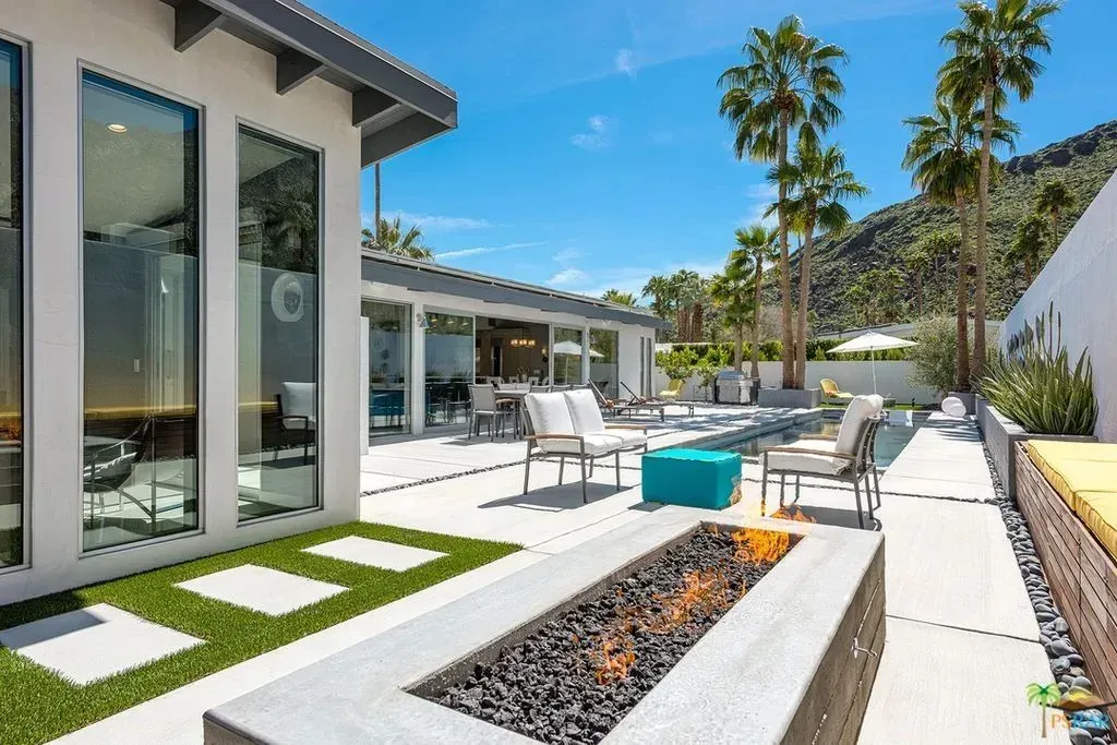 Modern patio with white lounge chairs, turquoise fire pit, and palm trees beside a sleek house