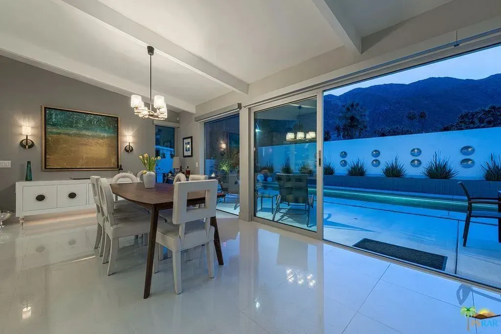 Modern dining room with glass doors opening to a blue-lit pool and mountain view at dusk