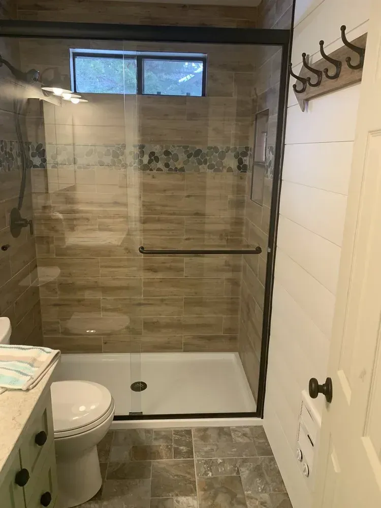 Bathroom with tiled walk-in shower, glass door, toilet, and vanity beside a window.