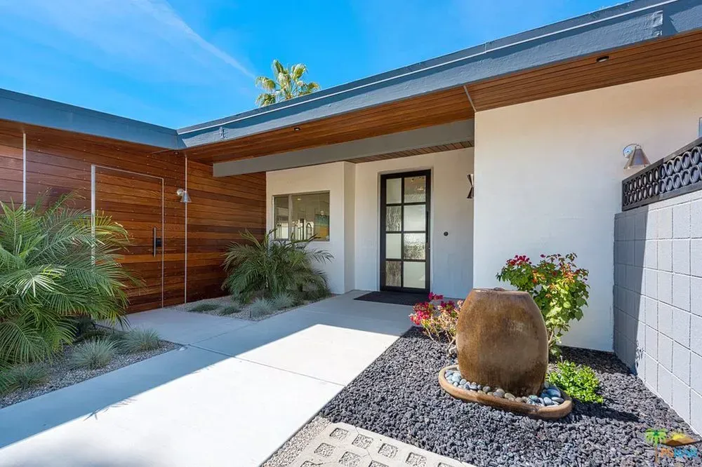 Modern courtyard entry with white stucco walls, wood siding, glass door, and a landscaped stone planter.