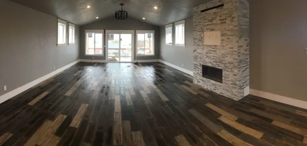 Empty modern living room with dark wood floors, gray walls, and a stone fireplace near large windows
