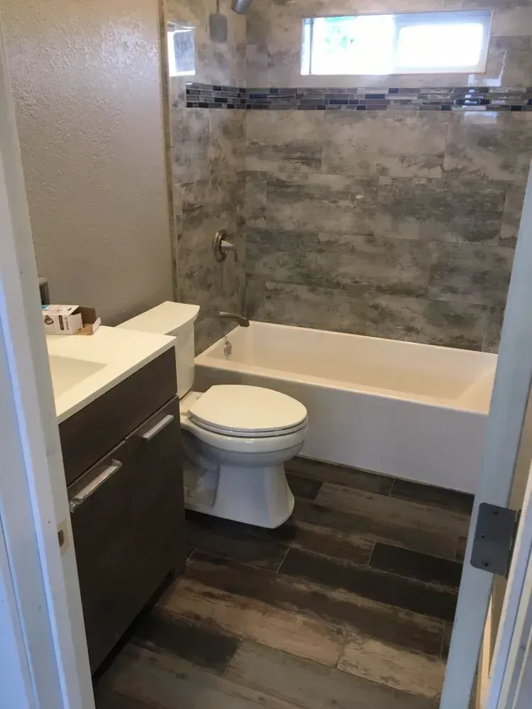 Small bathroom with toilet, vanity sink, bathtub, gray tile walls, and dark wood floor.