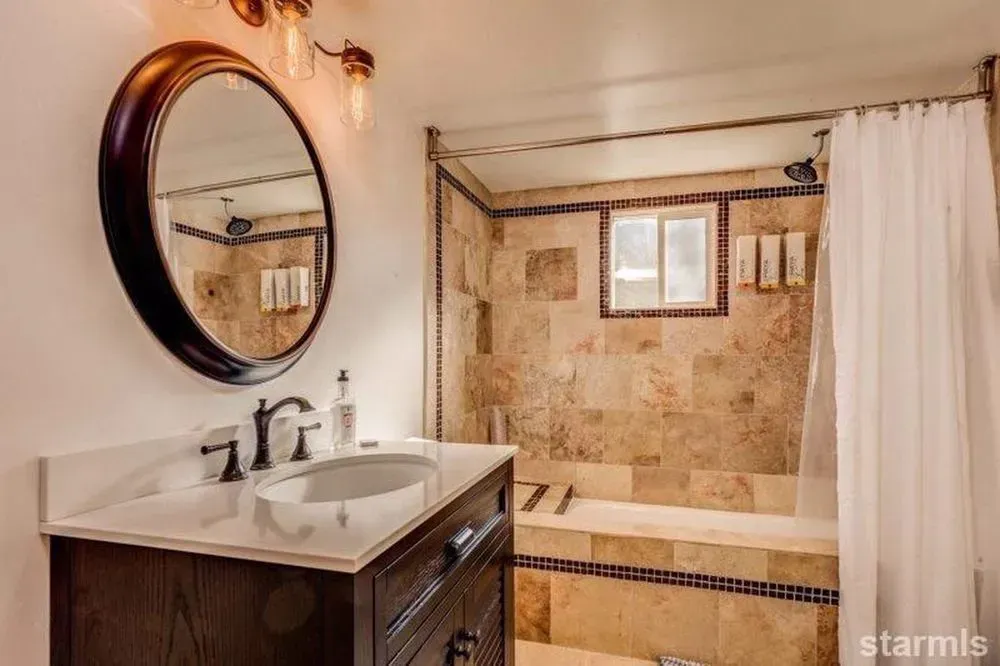 Bathroom vanity with round mirror beside a tiled shower and white curtain.