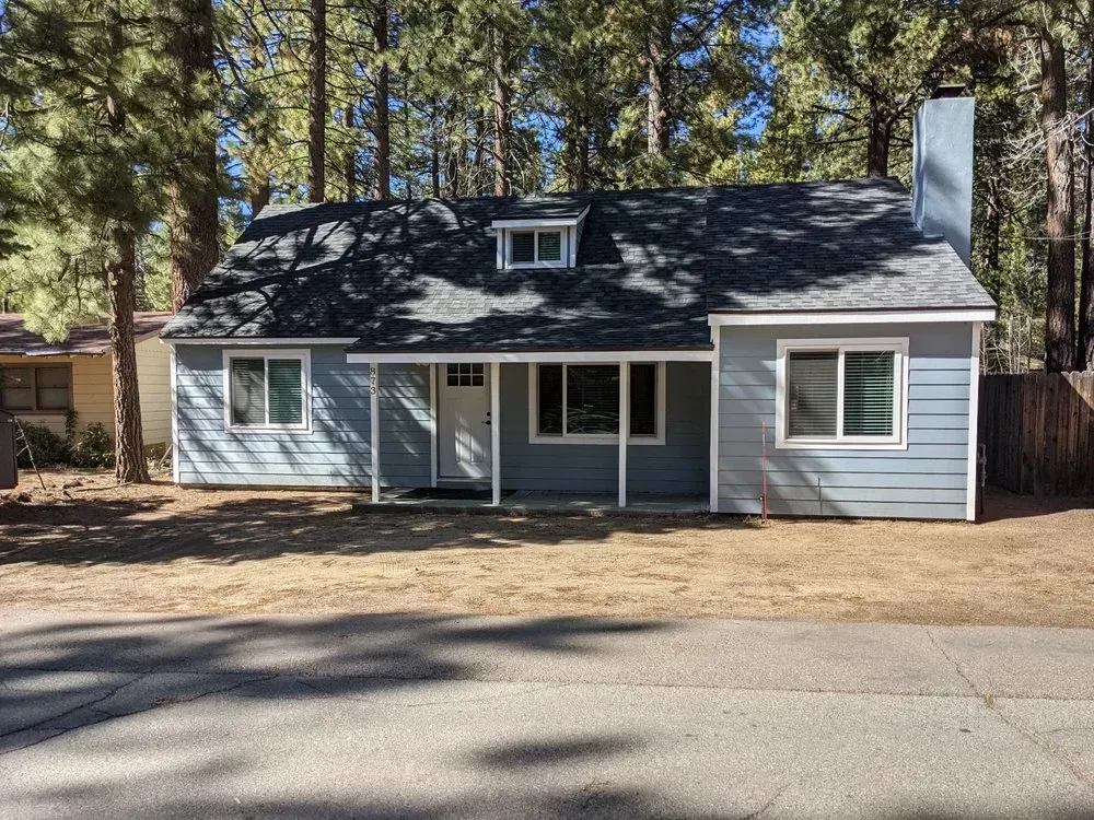Small blue house with dark roof and white trim, viewed from the street among tall trees.