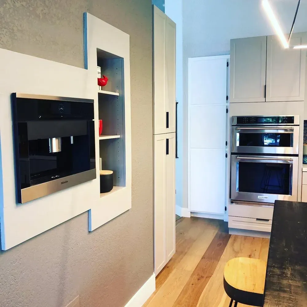 Modern kitchen with white cabinets, built-in oven, wall display shelves, and wood floor.