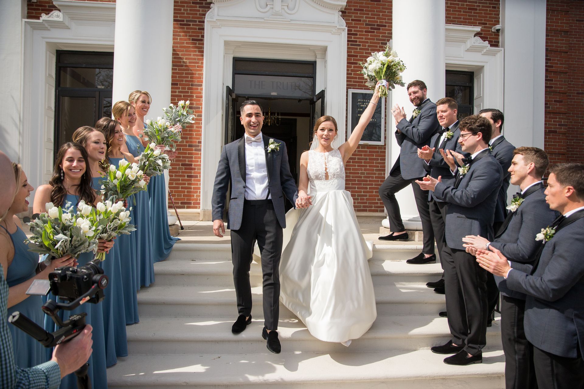 A bride and groom are walking out of a church with their wedding party taken by Ever After Studio.