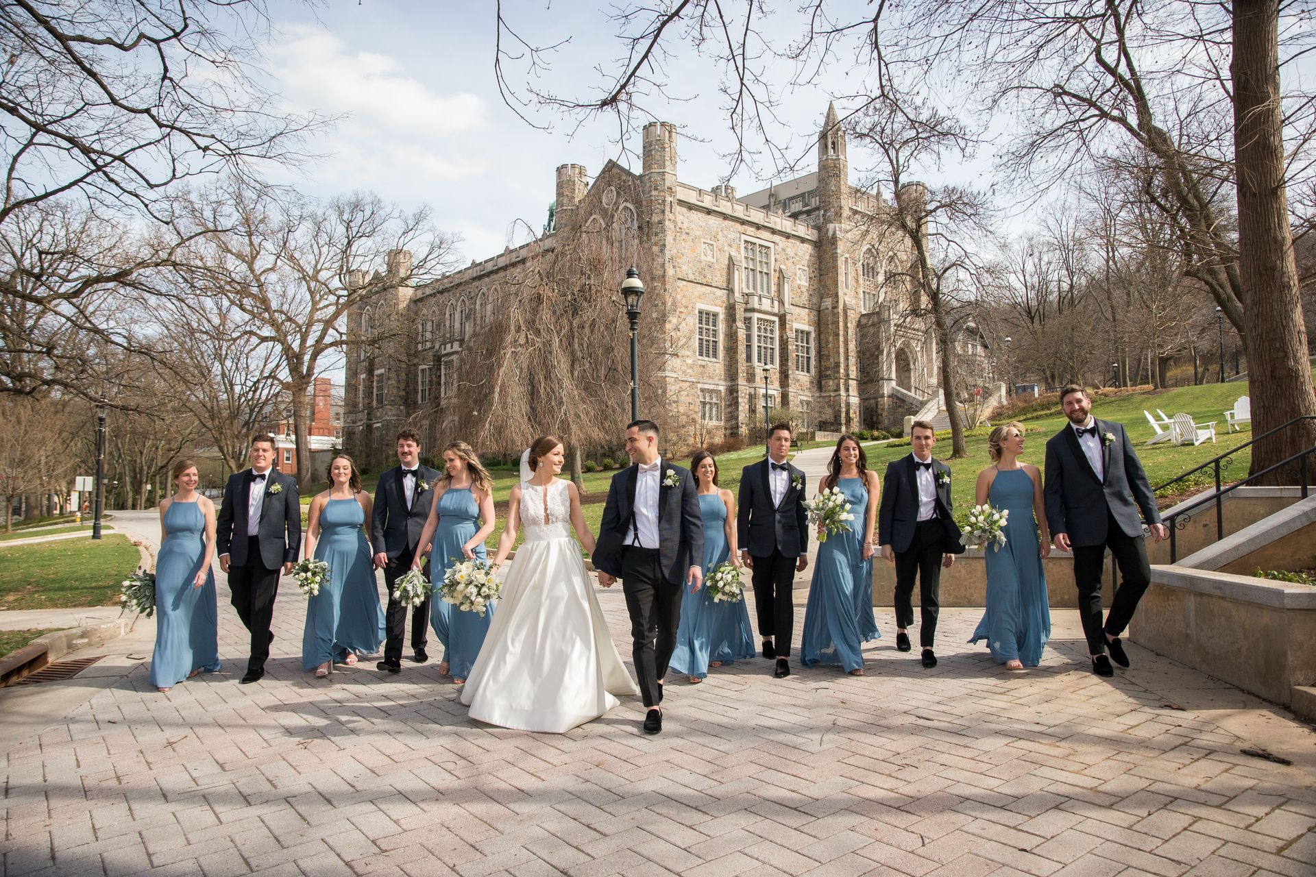 A bride and groom are walking with their wedding party in front of a large building taken by Ever After Studio.