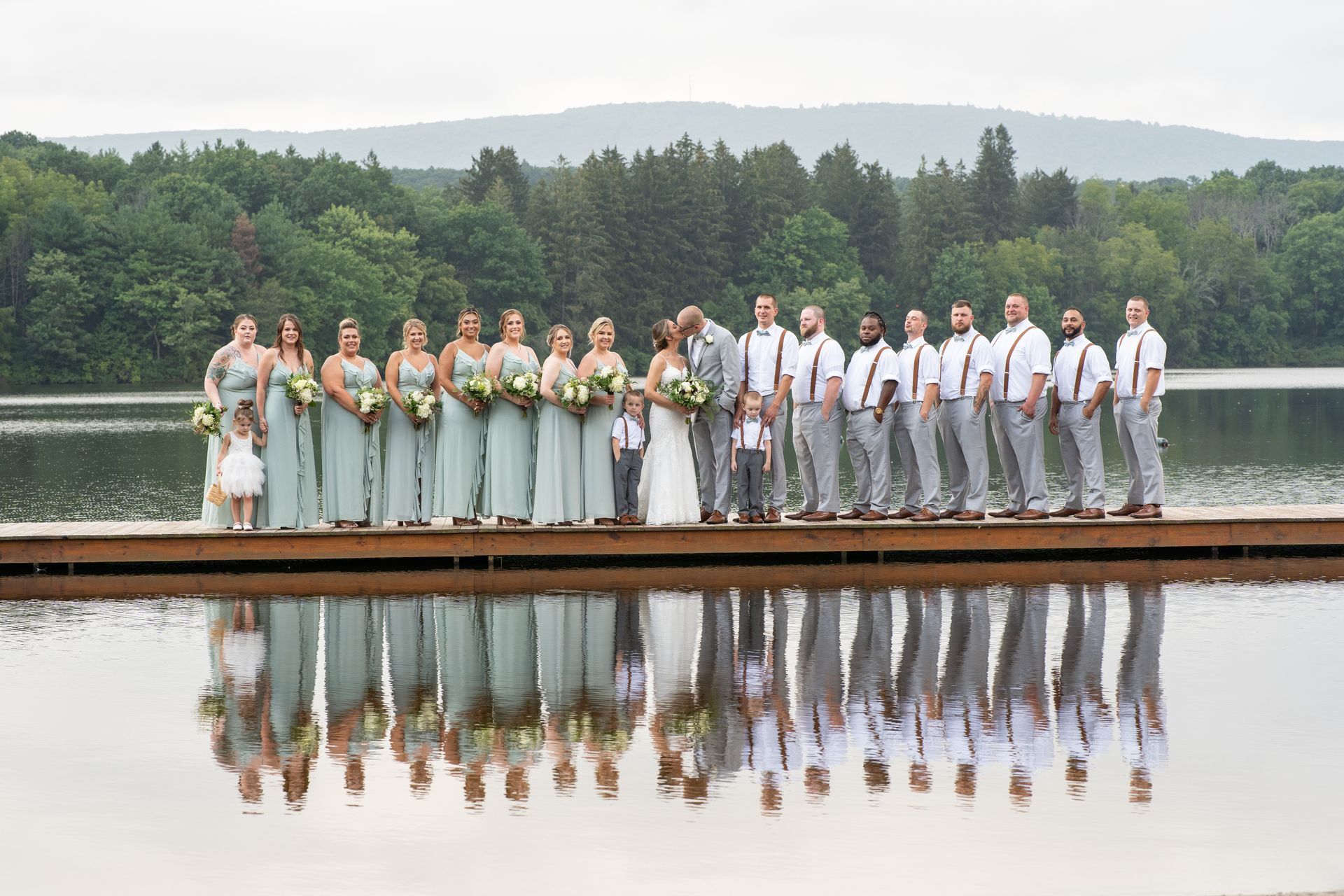 A wedding party is standing on a dock next to a lake taken by Ever After Studio.