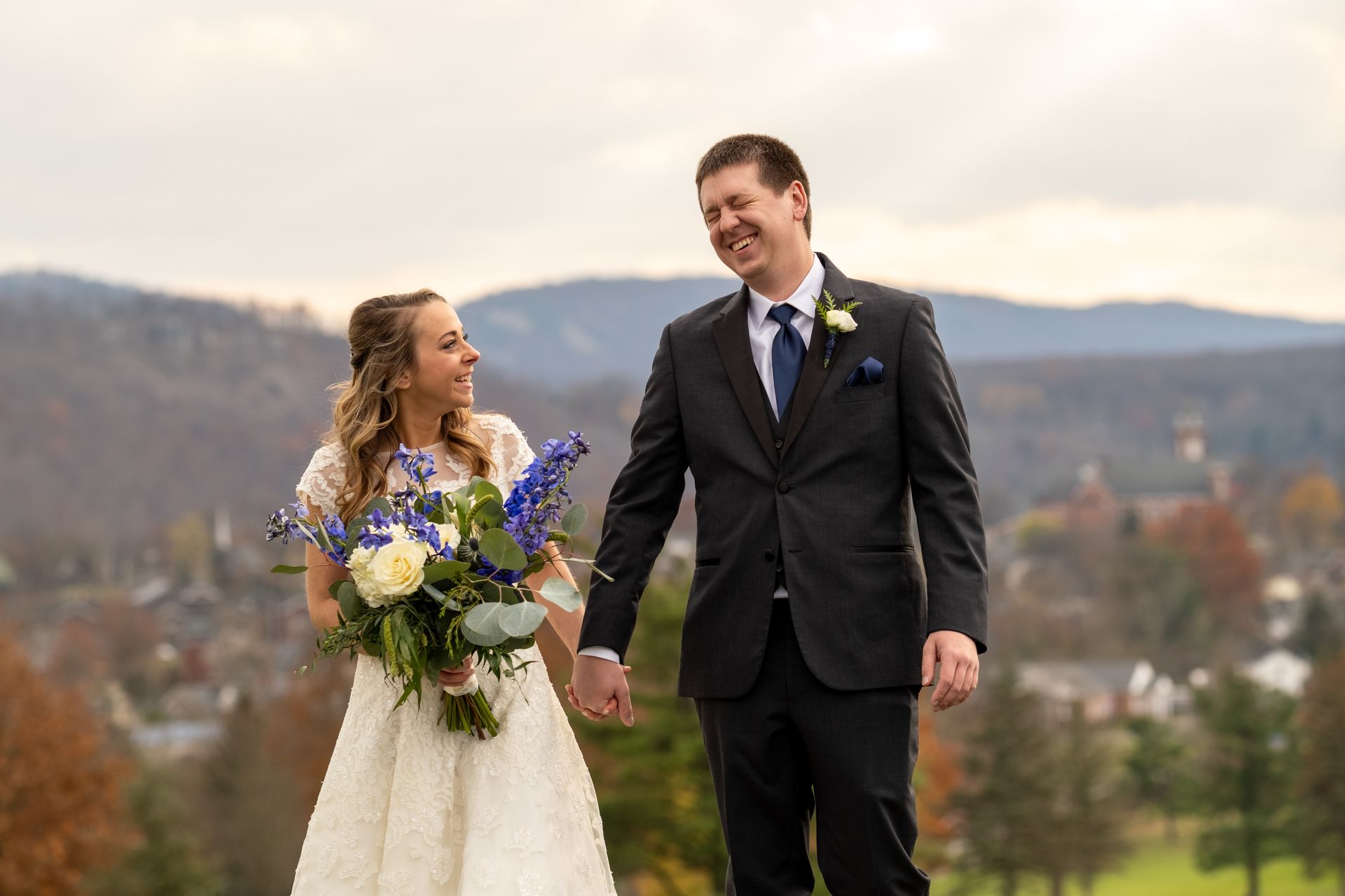 A bride and groom are holding hands and smiling while standing on top of a hill taken by Ever After Studio.