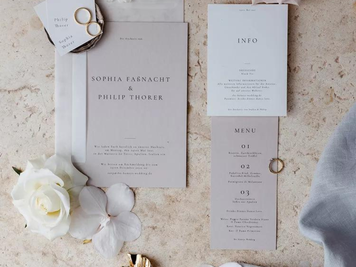 A wedding invitation is sitting on a table next to a white rose.