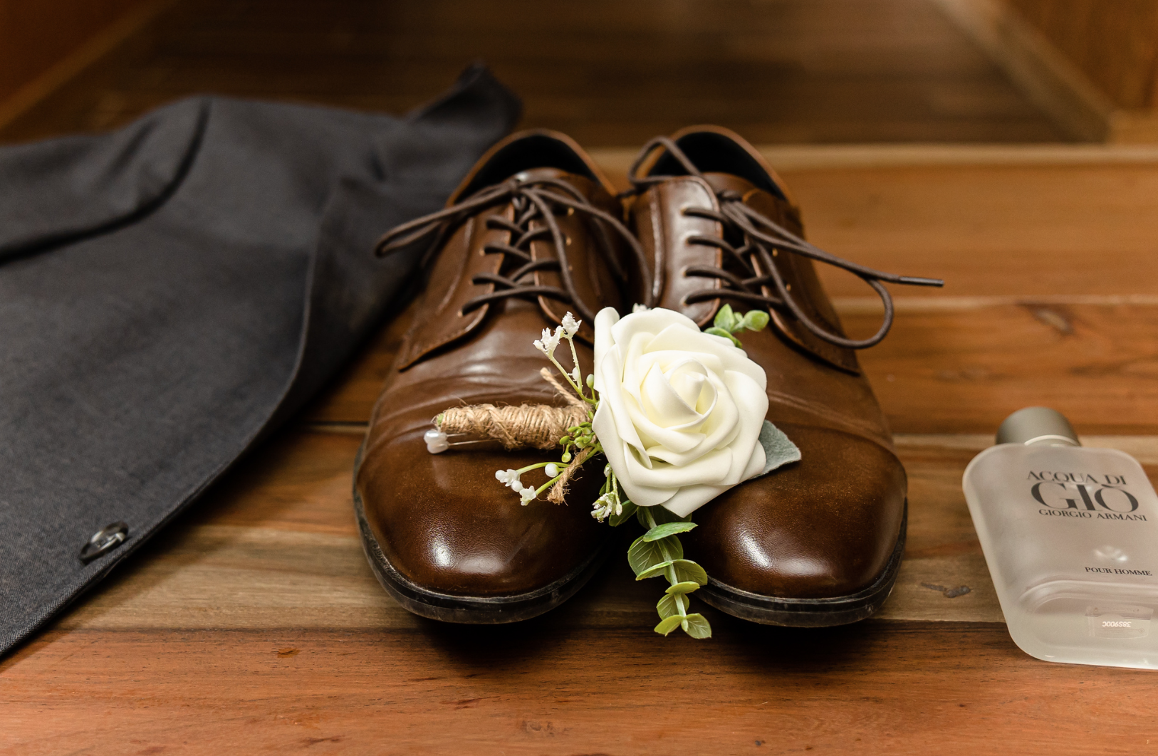 A pair of brown shoes with a white rose and a bottle of cologne on a wooden table taken by Ever After Studio.
