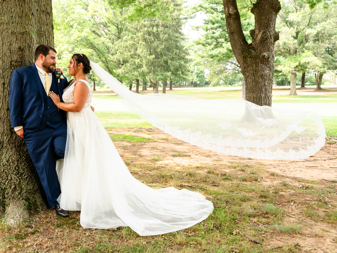 A bride and groom are leaning against a tree with their veil blowing in the wind by Ever After Studio