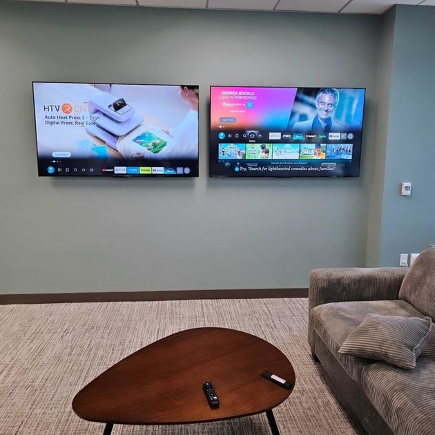 A room featuring two wall-mounted televisions, a coffee table with two remotes, and part of a grey couch.