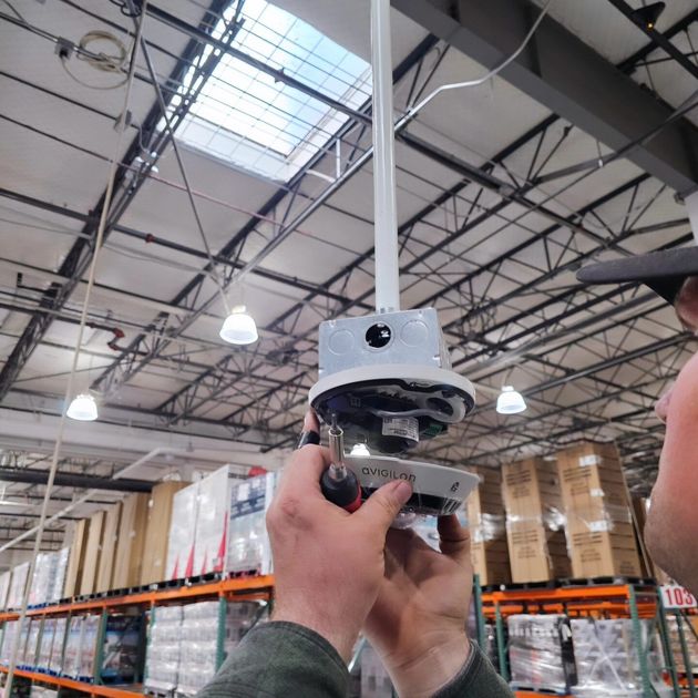A person holds and repairs a ceiling-mounted light fixture inside a large warehouse with stacked pallets.