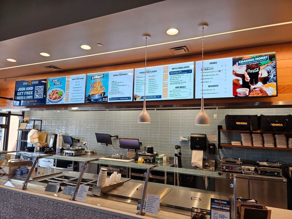 A brightly lit restaurant counter featuring multiple digital menu screens mounted above a clean, stainless steel prep area.