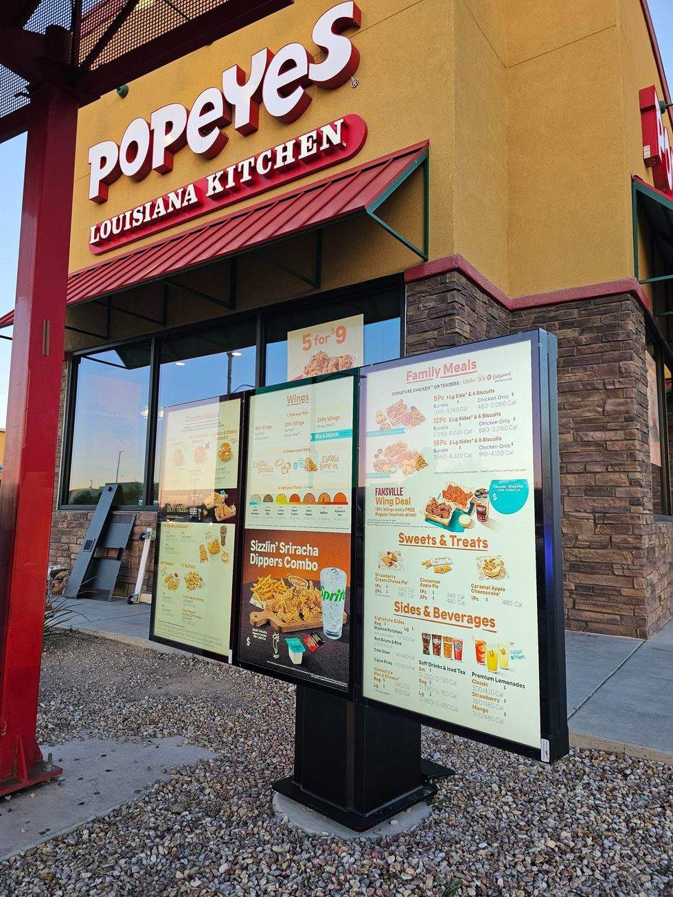 An outdoor menu board stands in front of a Popeyes Louisiana Kitchen restaurant building with a red metal support beam.