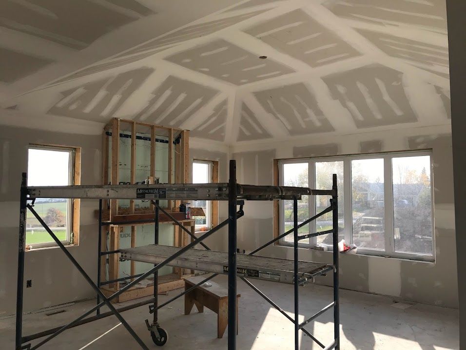 Interior room under renovation with scaffolding, unfinished drywall ceiling, and large windows.