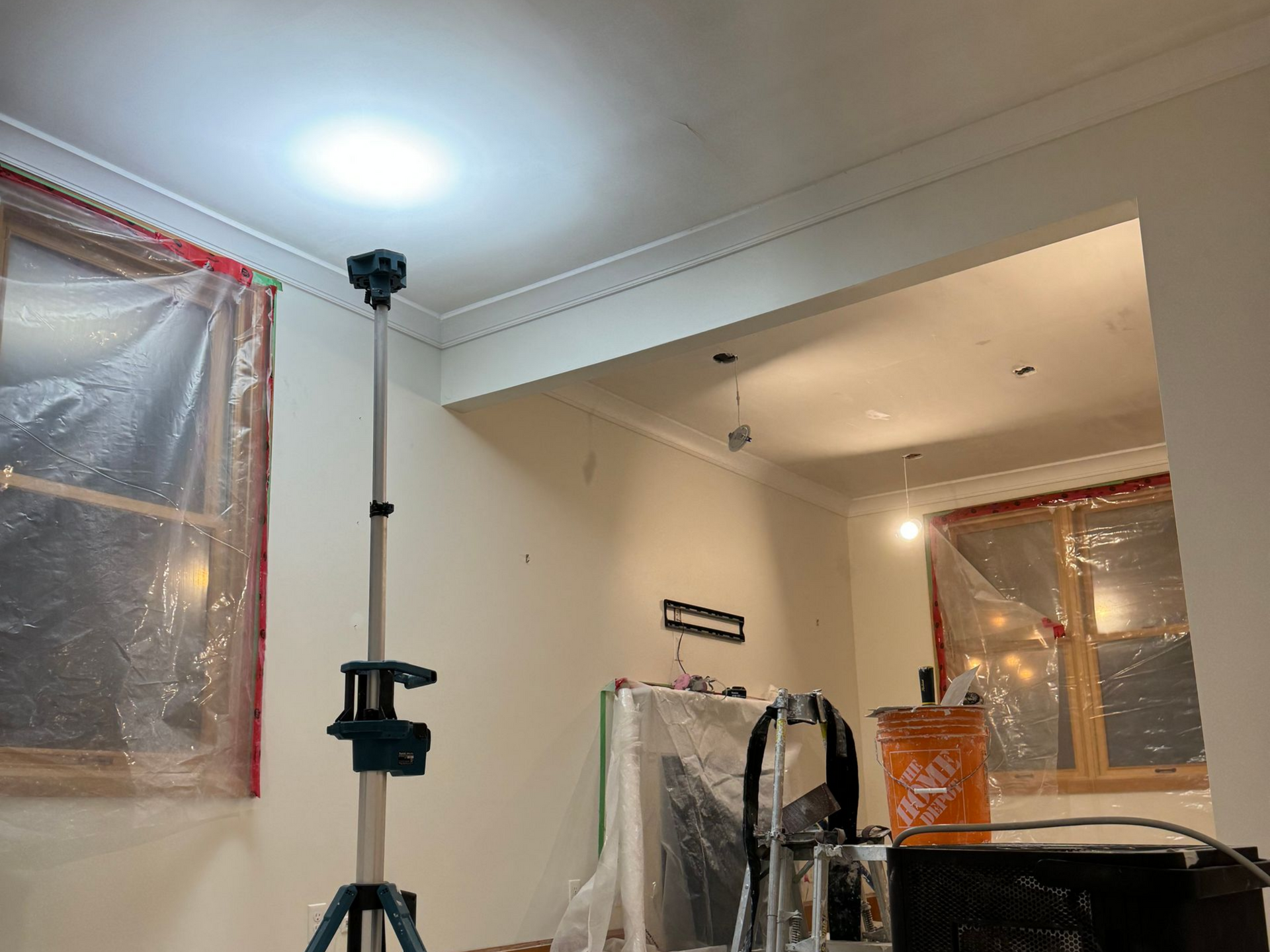Renovated room with a work light on a tripod; plastic covers windows. White walls and crown molding.