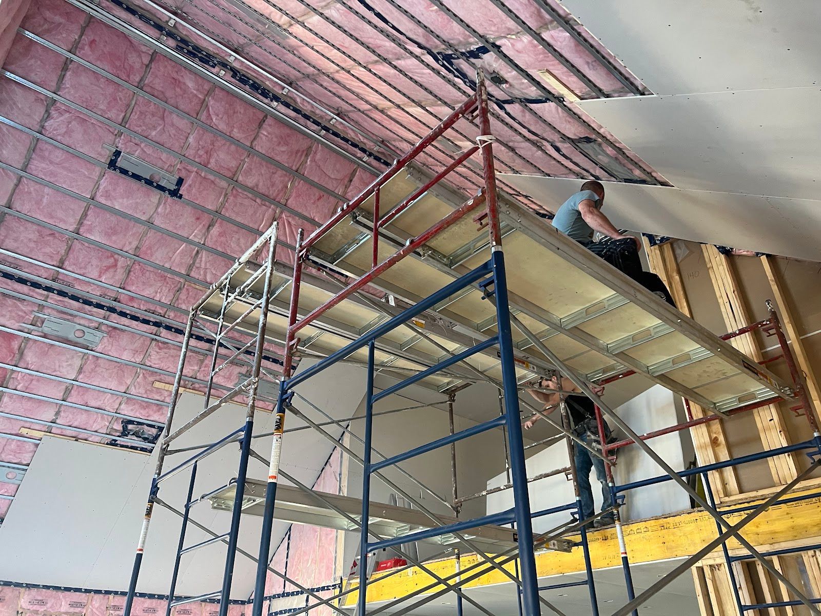 Workers on scaffolding installing a ceiling in a room with exposed insulation and drywall.