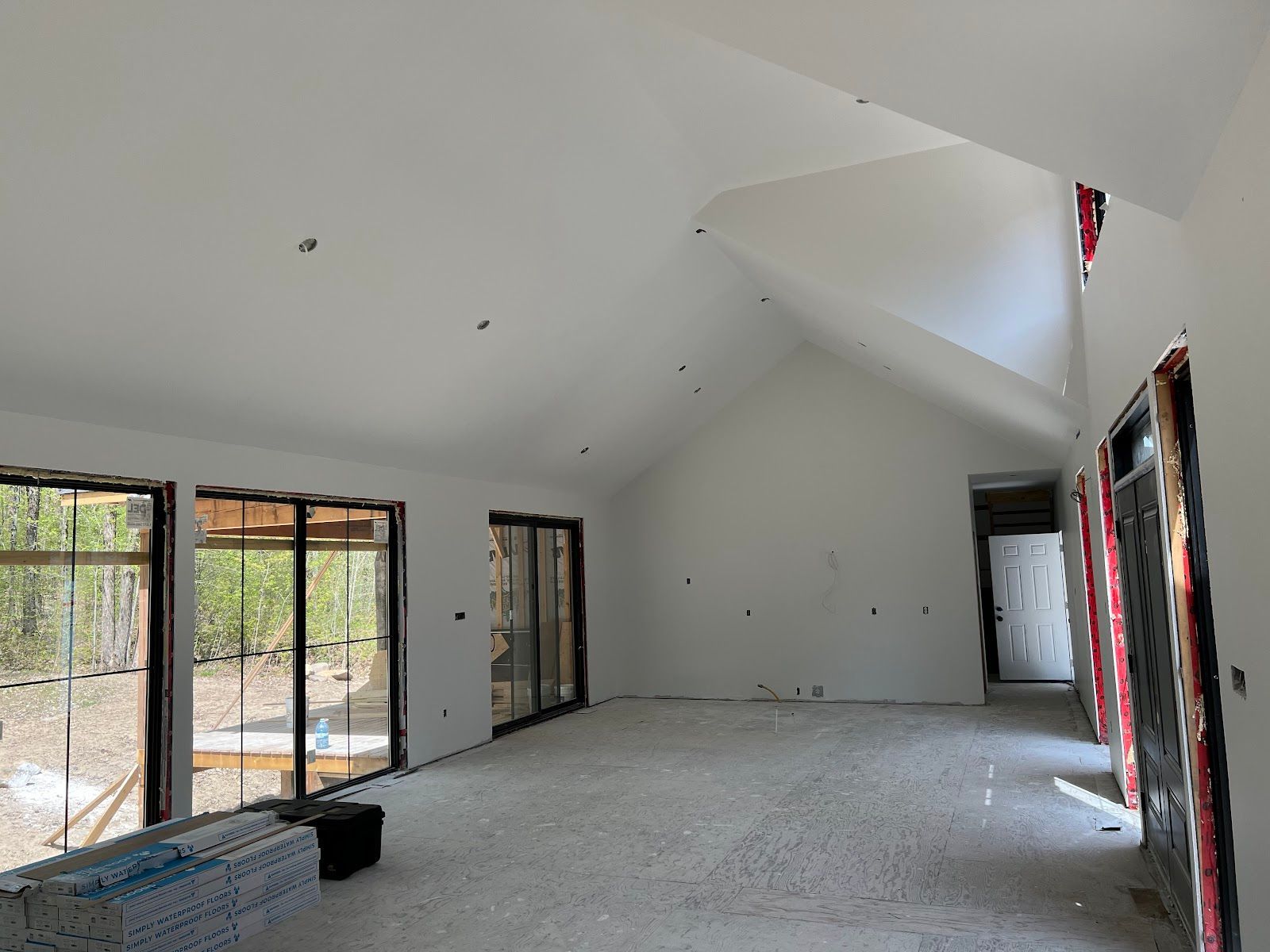 Empty unfinished living room with tall white vaulted ceiling, sliding glass doors, and bare concrete floor.