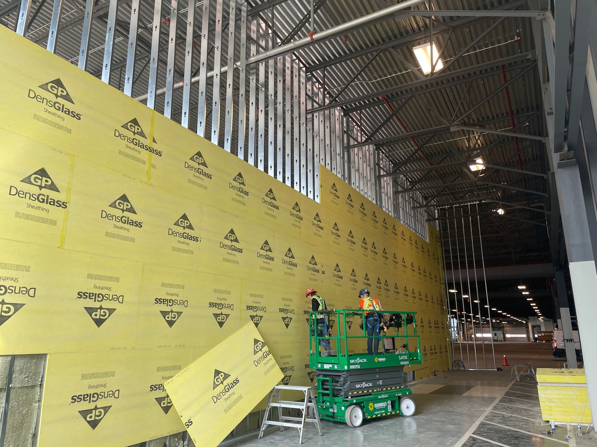 Workers installing yellow insulation on a metal-framed wall inside a large construction site.