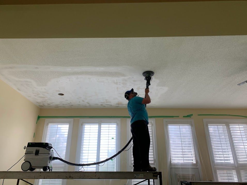Man sanding a textured ceiling, using a dust collection system. Indoors with windows and scaffolding.