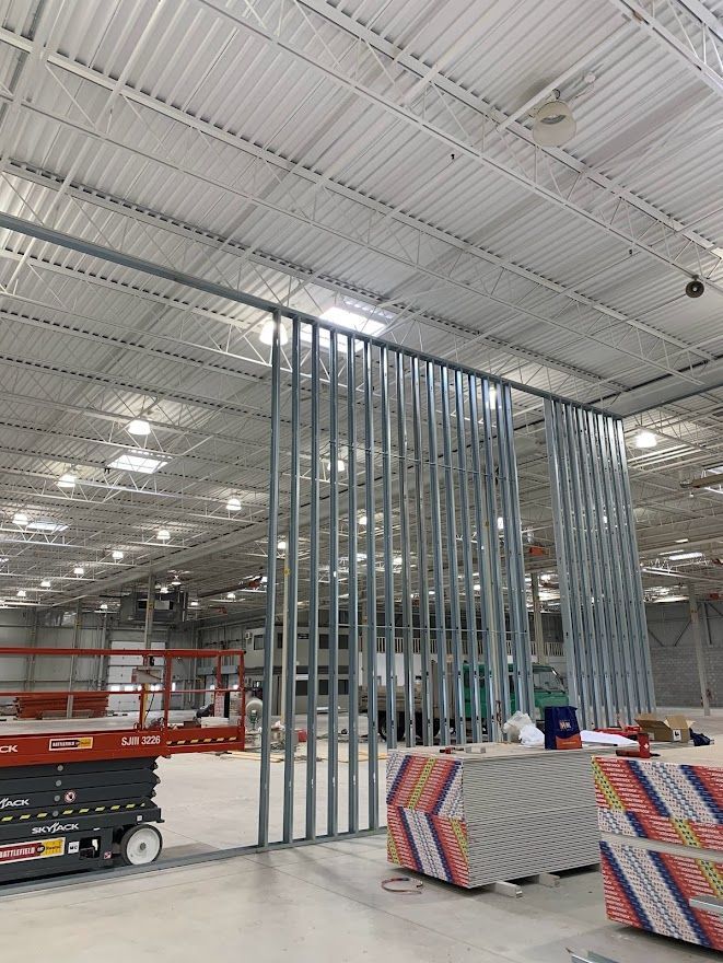 Interior view of a building under construction. Metal studs form a partial wall; drywall stacks are on the floor.