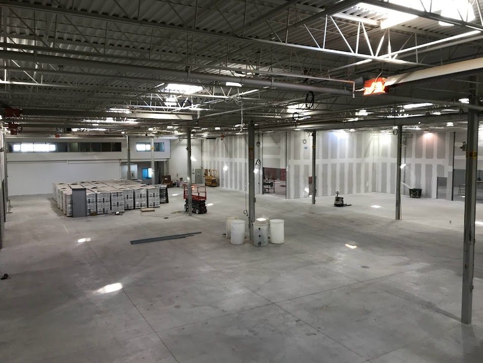 Empty industrial warehouse with concrete floor, exposed ceiling beams, and stacks of materials near the back.