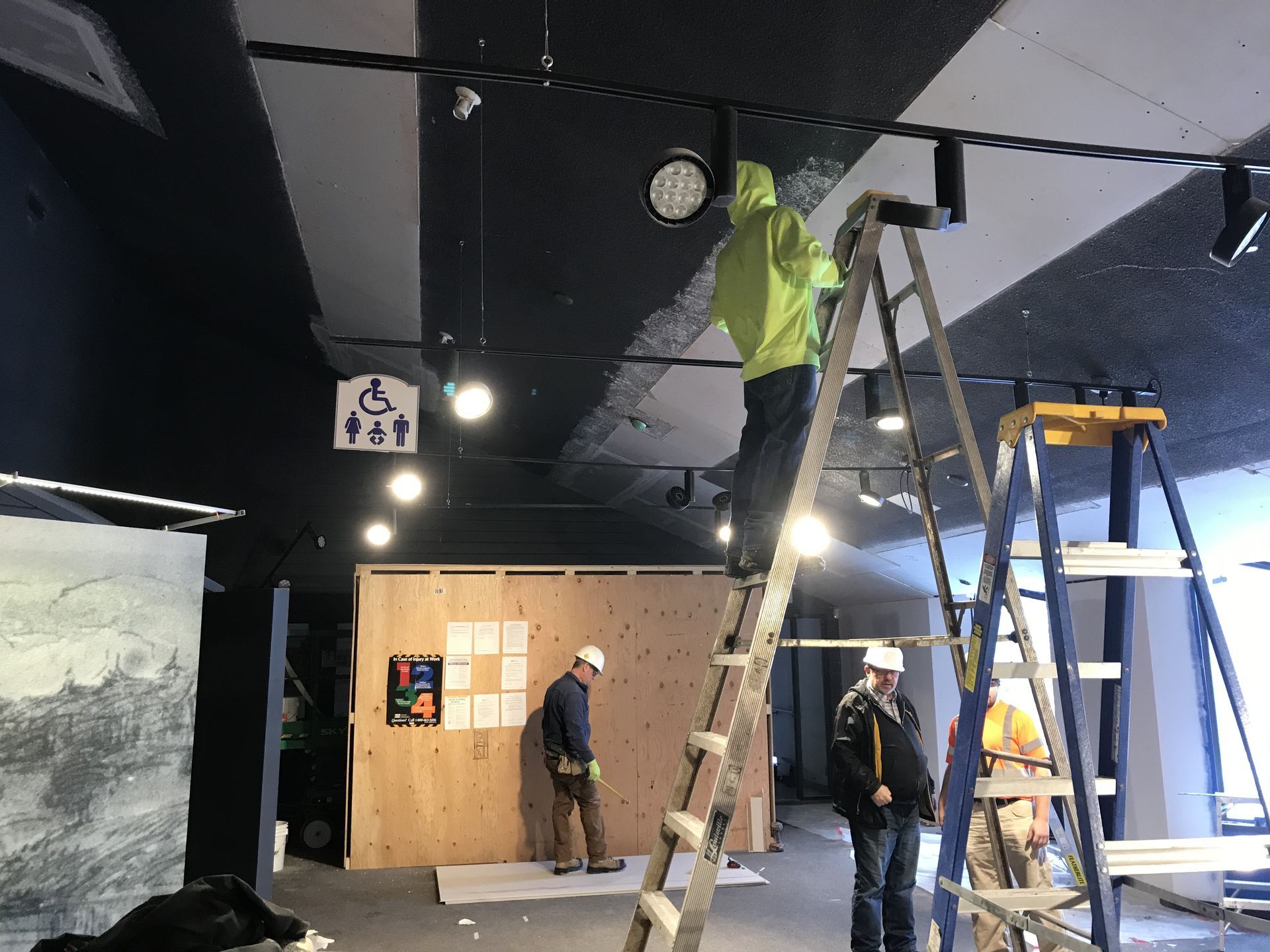 A man in a green jacket is standing on a ladder in a room.