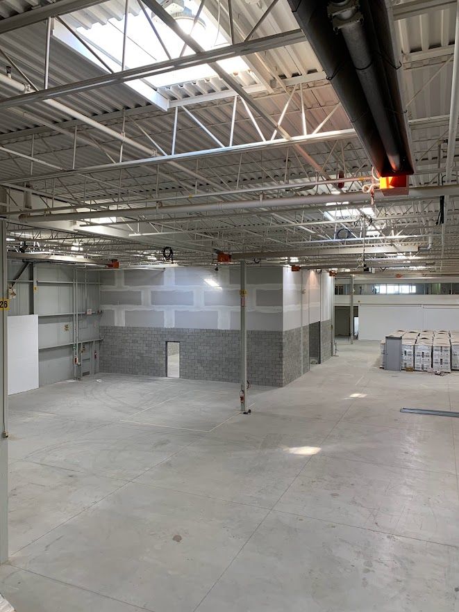 Interior view of a large, unfinished warehouse space with concrete floors, exposed ceiling beams, and partially built walls.