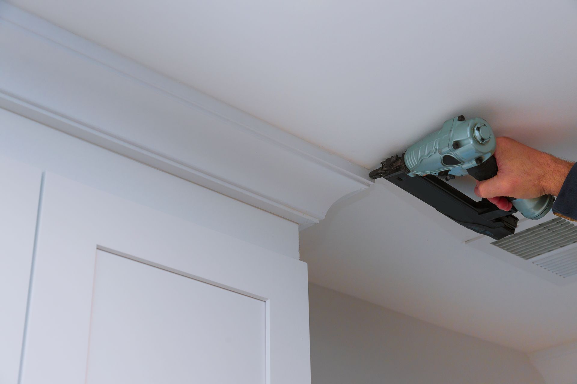 A person using a nail gun to install crown molding on a white ceiling above a cabinet.