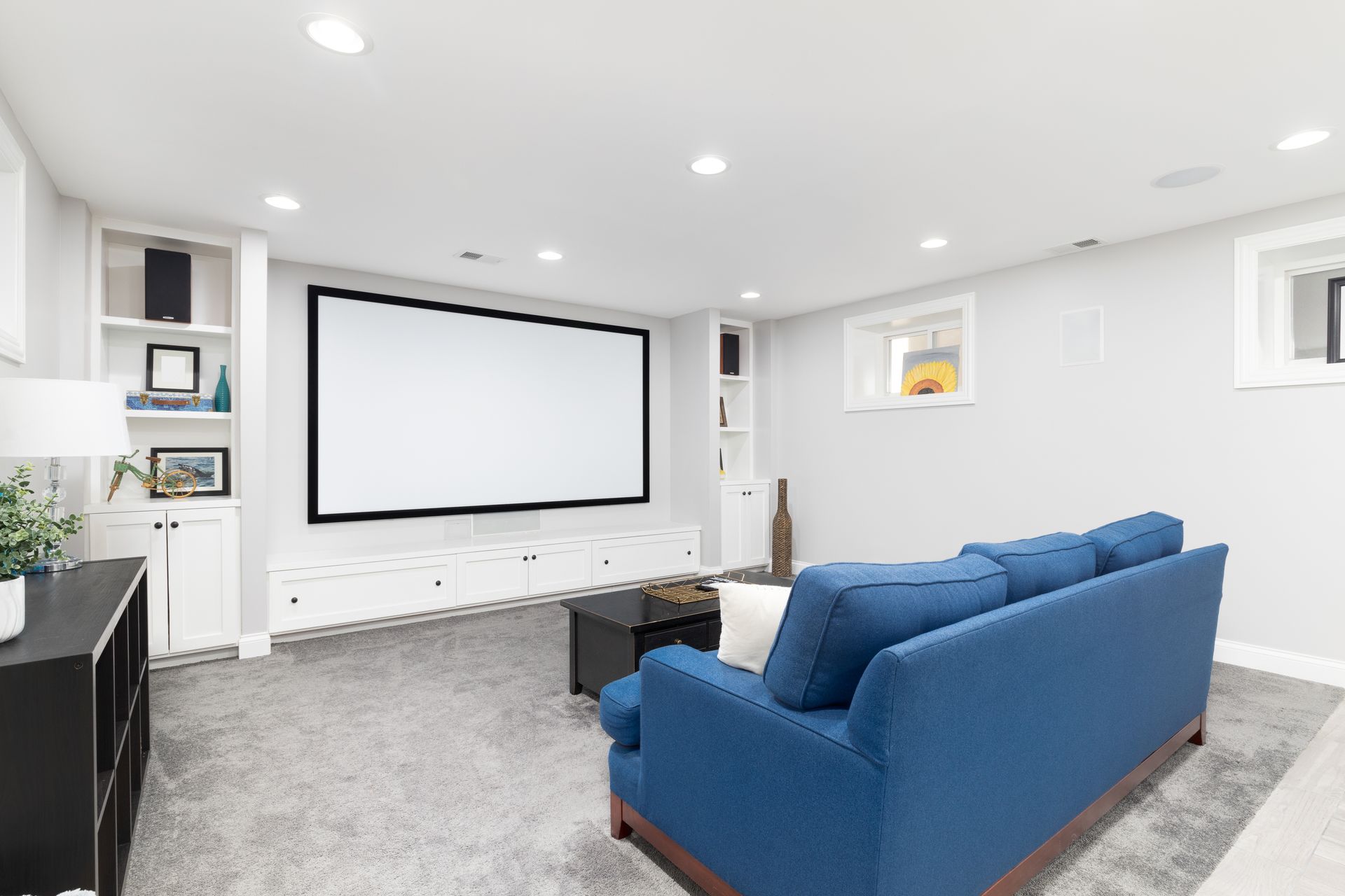 Basement living room with blue couch, large projection screen, gray carpet, and white built-ins.
