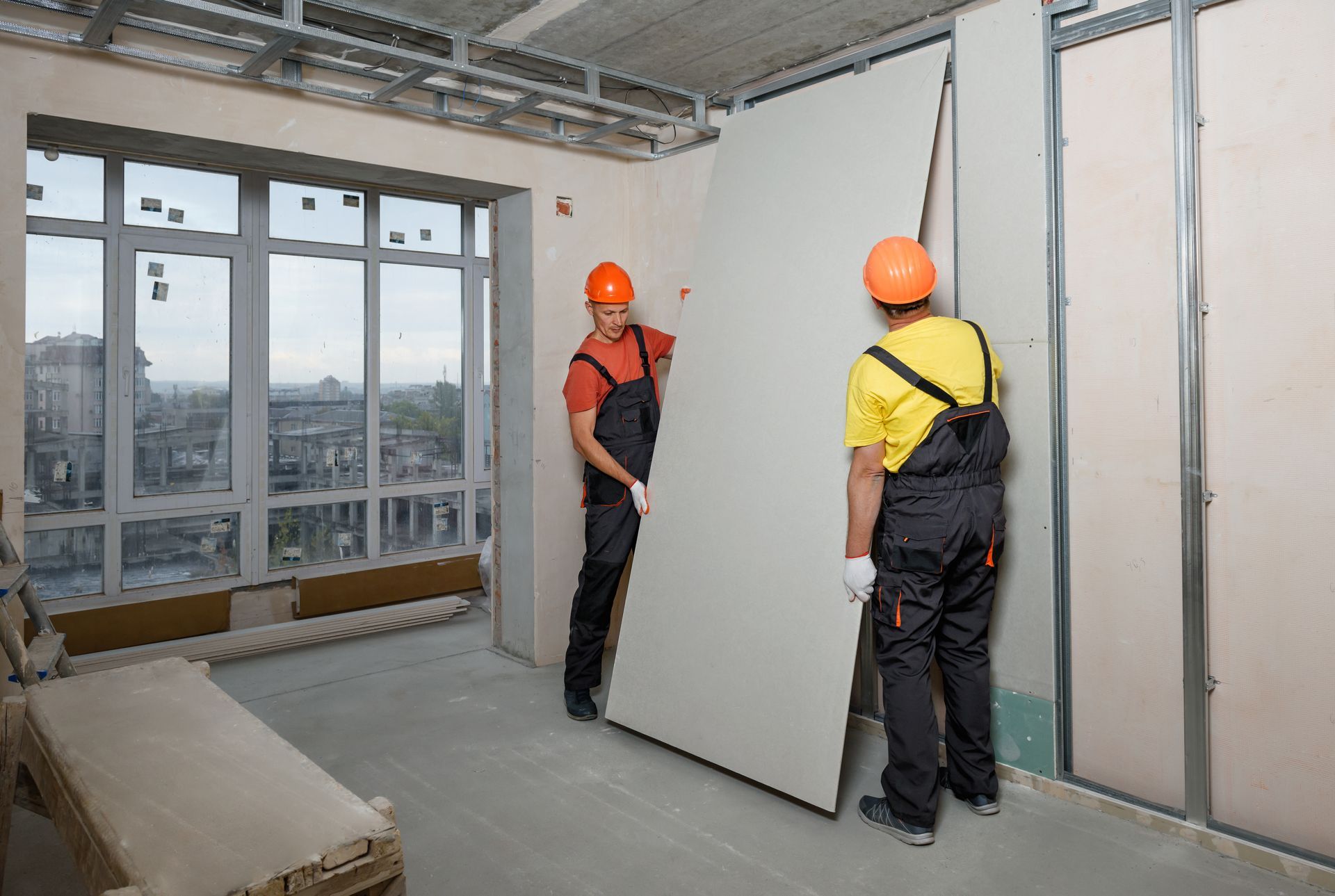 Two men are carrying a large piece of drywall in a room.