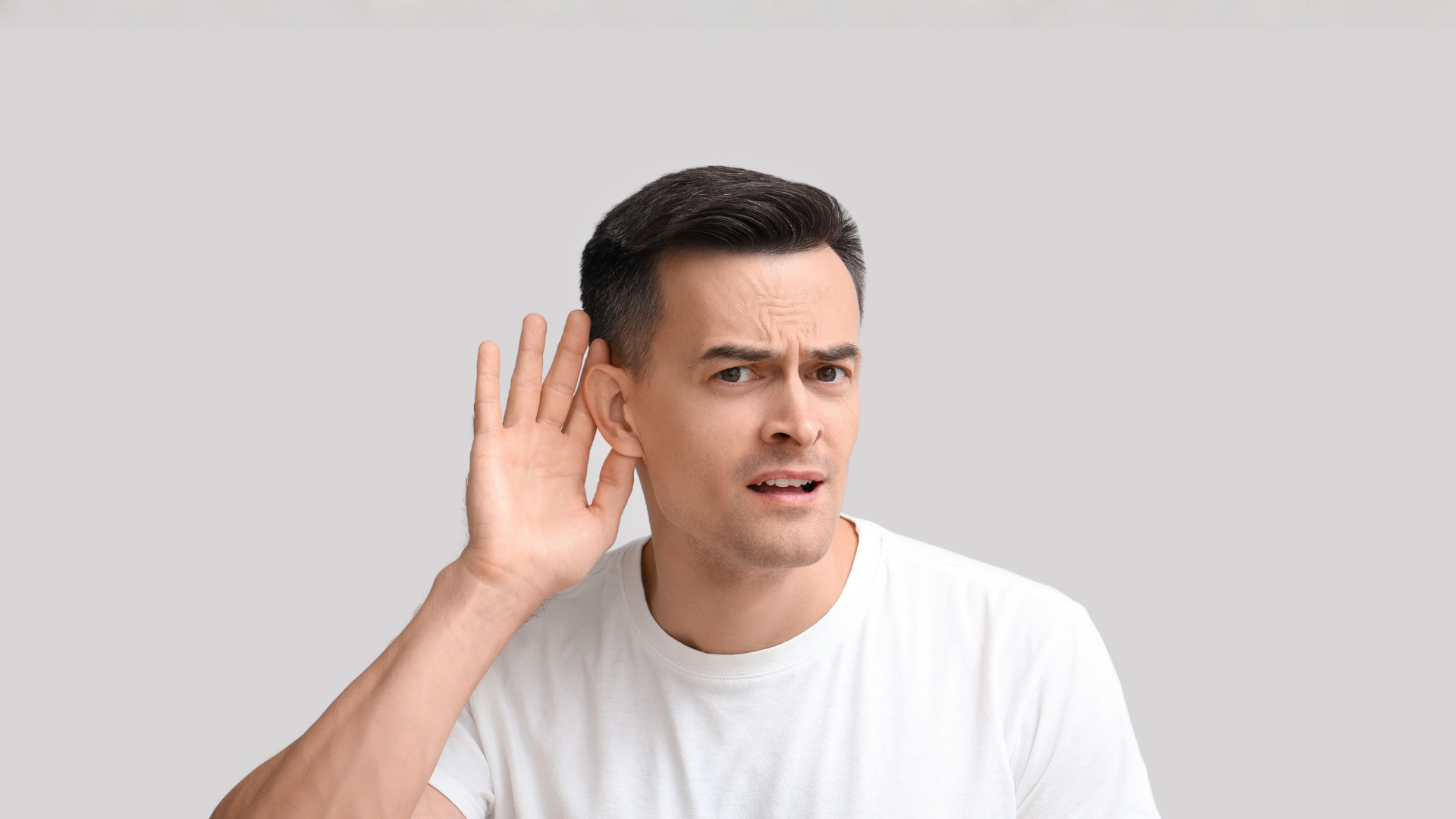 Man in white shirt, hand cupped behind his ear, looking with a confused expression.