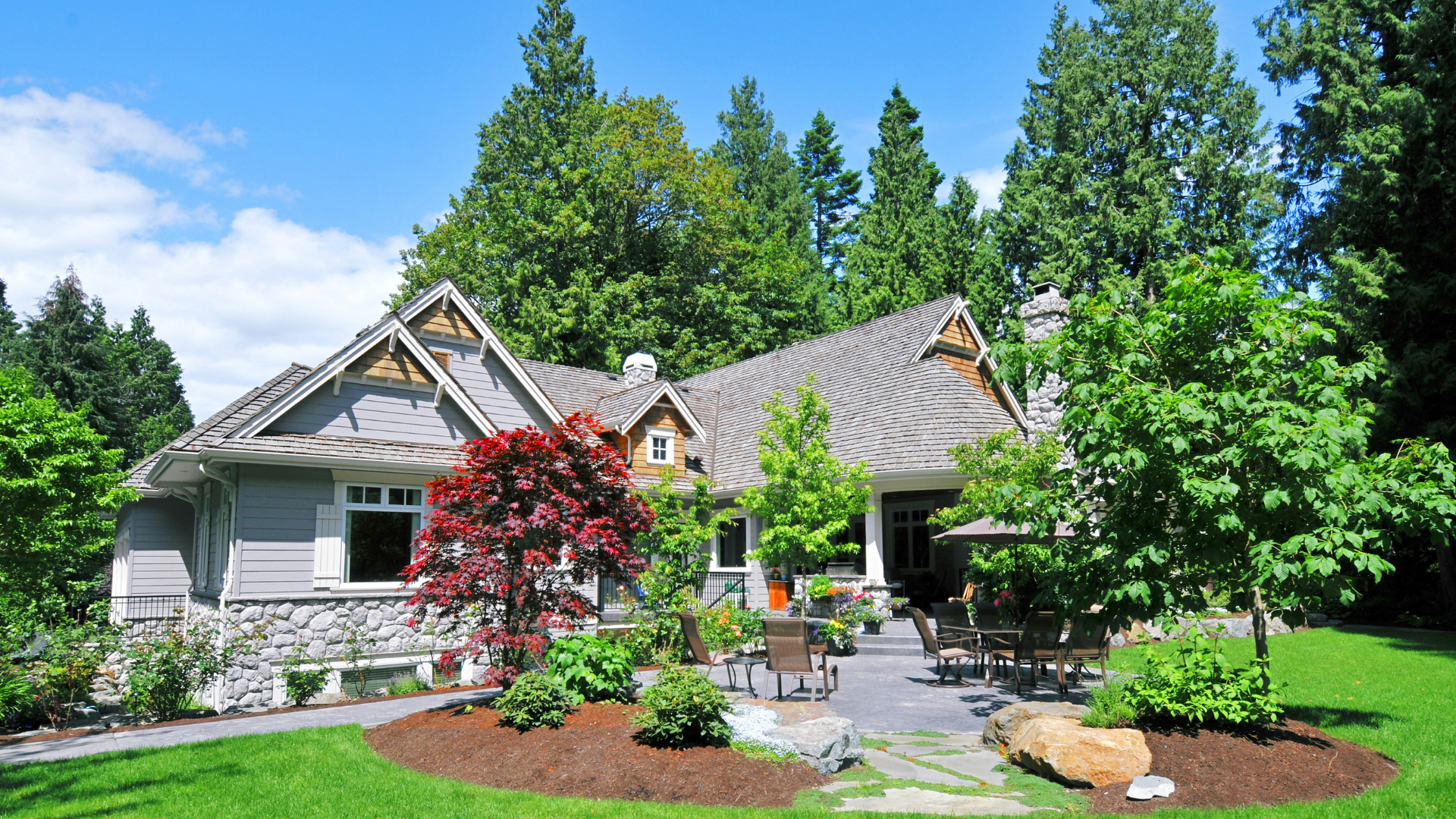 A grey craftsman-style house with stone accents surrounded by trees, a lush lawn, and a landscaped stone patio.