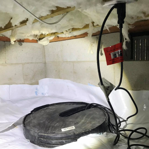 A black sump pump and white container are connected to an electrical outlet in a crawl space.