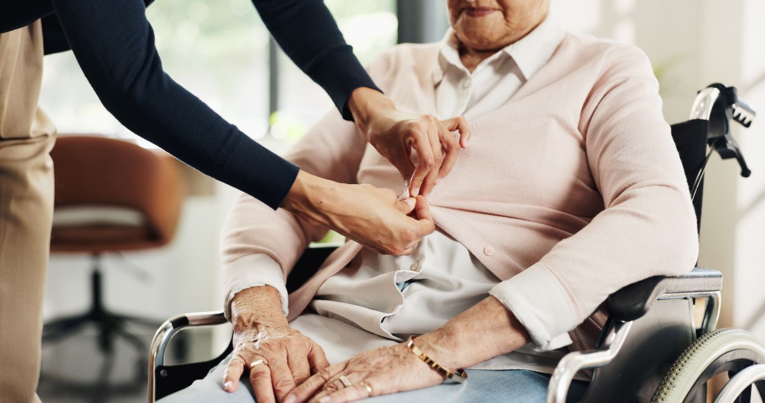 Caregiver adjusting a seated patient’s shirt in a wheelchair, with a cane nearby. Caregiver adjusting a seated patient’s shirt in a wheelchair, with a cane nearby.
