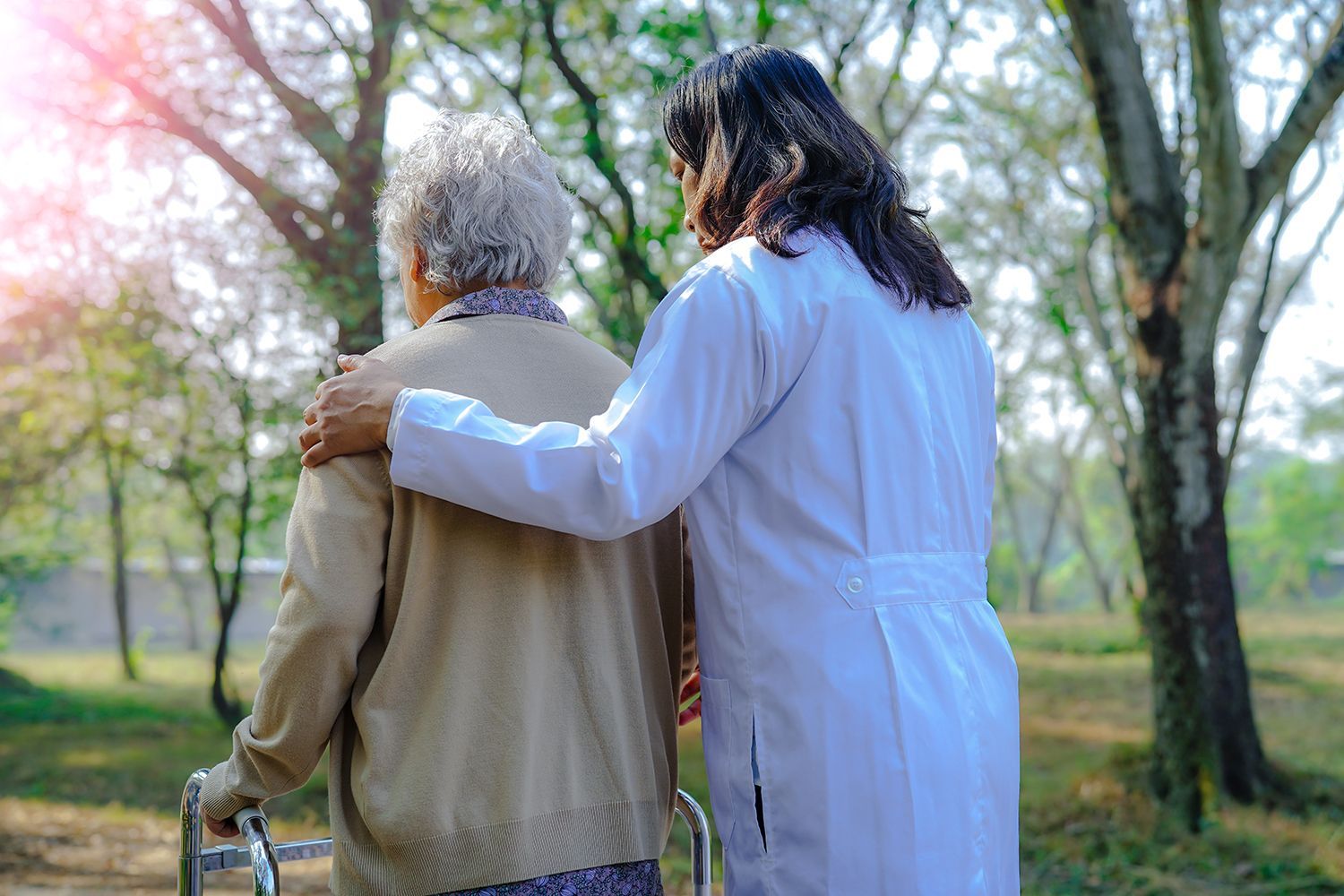 Two people walking in a park, one in a white coat with an arm around the other