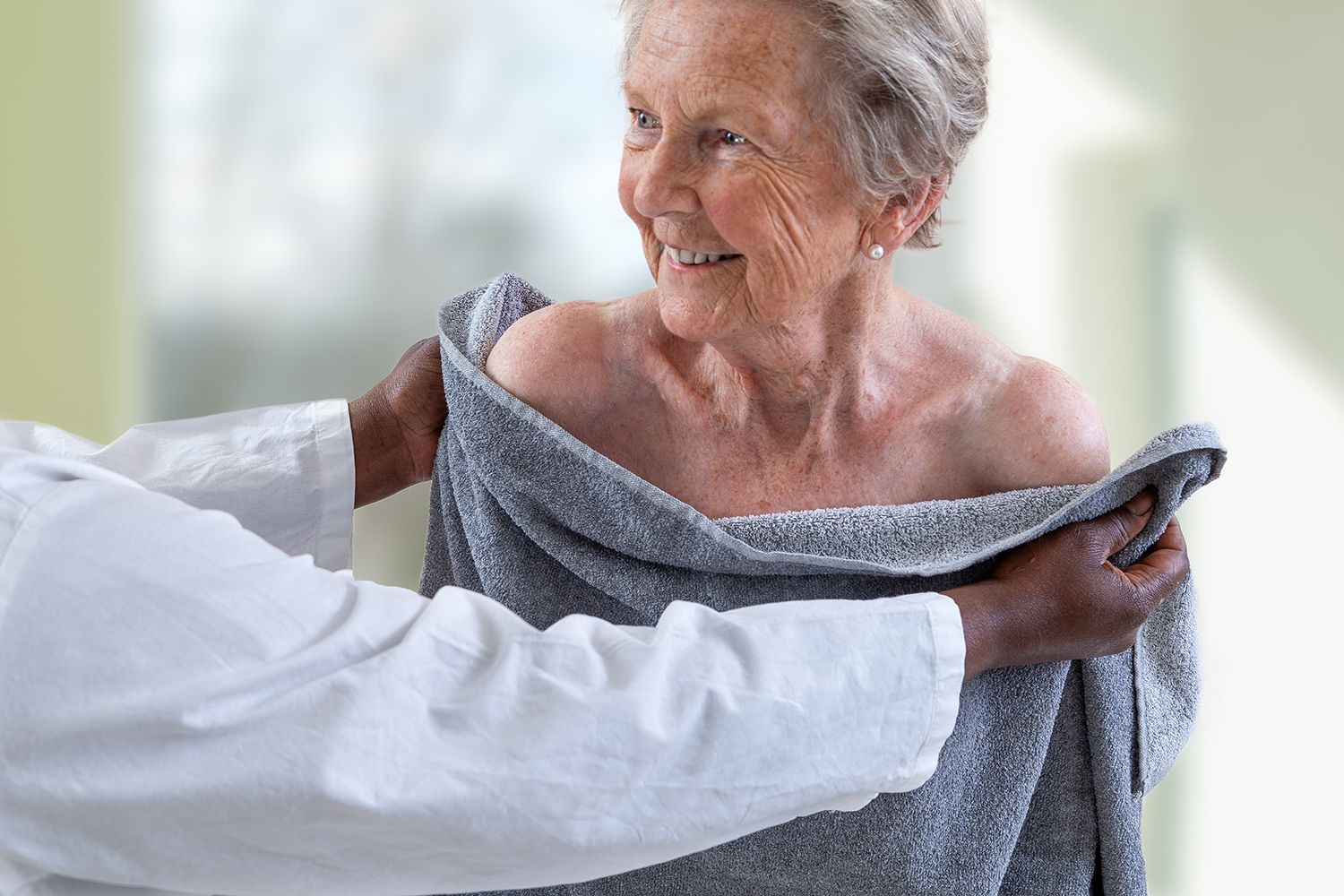 Caregiver helps a smiling patient put on a gray sweater in a bright room