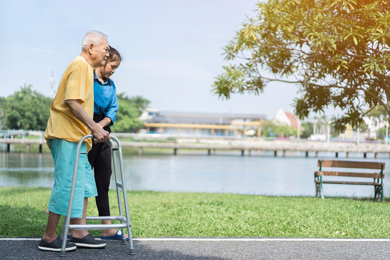Elderly couple walking with a walker by a lakeside path, with a bench and tree nearby.
