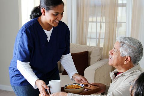 Caregiver serving a meal to an elderly man in a living room, both smiling.