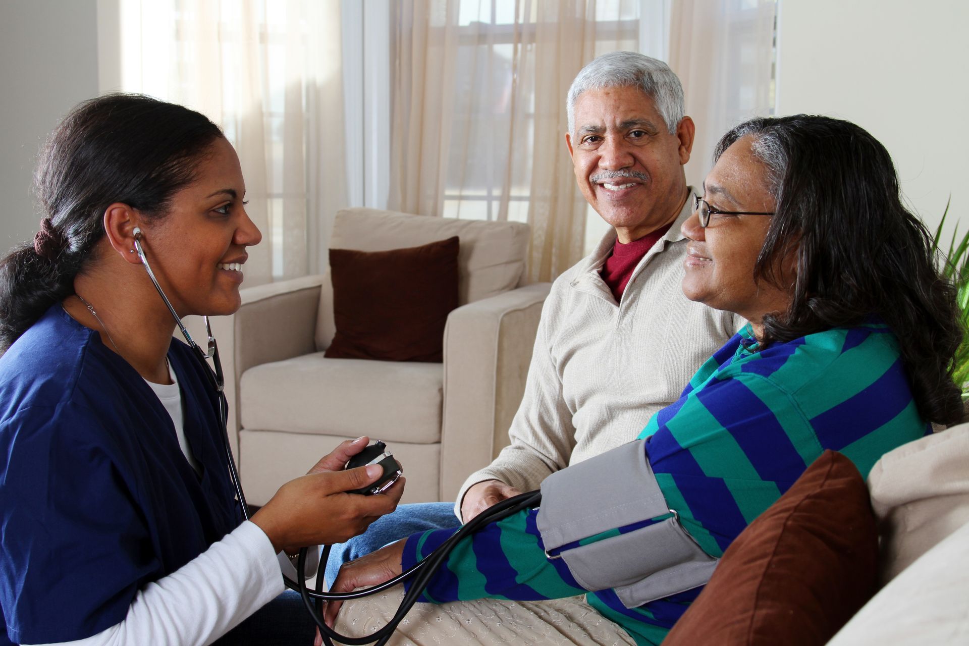 Healthcare worker checking a patient’s blood pressure while another adult sits nearby in a living room