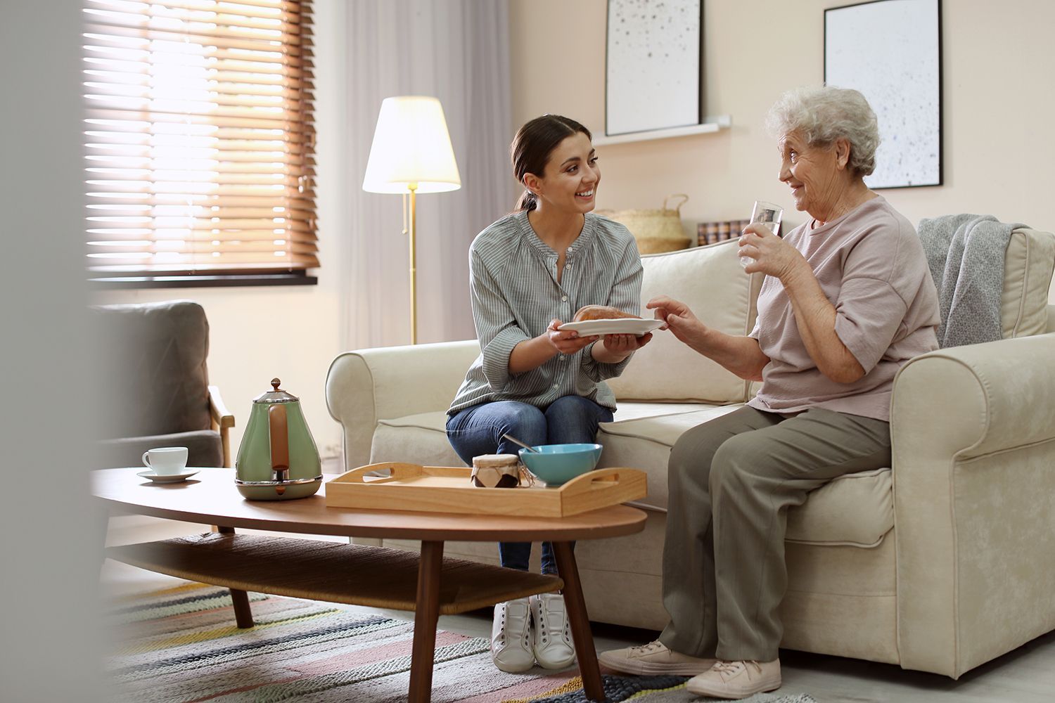 Two people talking on a beige sofa in a bright living room with a coffee table and lamp. Two people talking on a beige sofa in a bright living room with a coffee table and lamp.